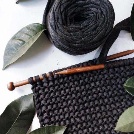 a piece of knitting in progress. Wooden straight needles in a black garter stitch fabric made from tshirt yarn. The piece is styled with dark green leaves.