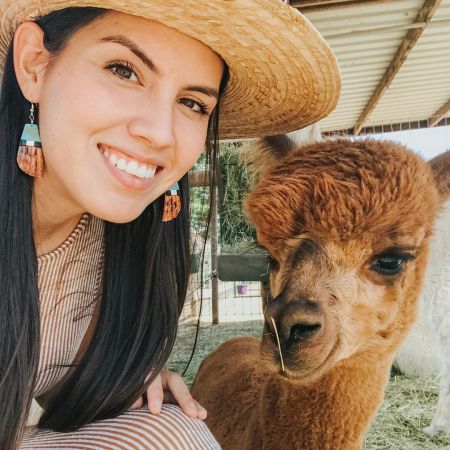 Jen, an Indigenous woman with long dark hair, wearing a straw hat and a brown striped dress, smiles brightly into the camera. She’s kneeling in a shed in an outdoor area with grass on the ground. Next to her is a reddish brown alpaca with a piece of hay sticking out of its mouth.